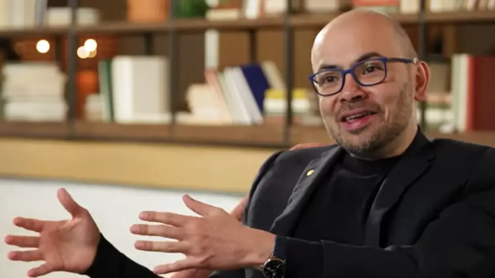 Demis Hassabis, CEO of Google DeepMind, speaking in an interview with hands gesturing, seated in a cozy room with bookshelves in the background.
