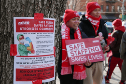 Nurses wearing red strike apparel hold signs reading “Safe Staffing Saves Lives” during a nurse strike, with a strike flyer taped to a tree in the foreground.