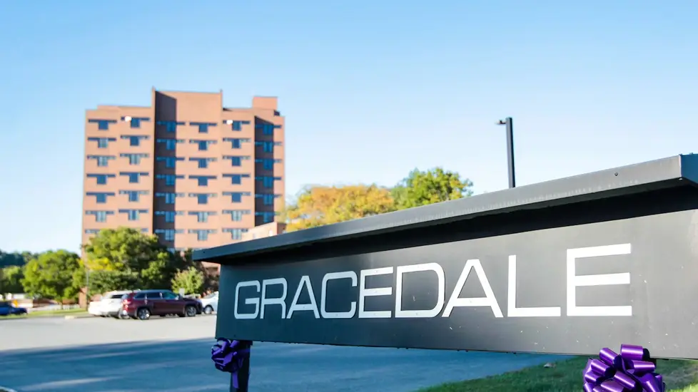 A large, multi-story brick building stands in the background, with a parking lot and several cars in front. In the foreground, a black sign with bold white letters reads "GRACEDALE"
