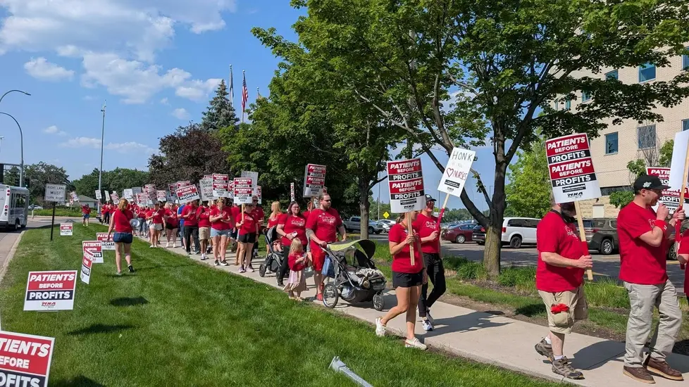 A large group of nurses and supporters march along a sidewalk outside a hospital during a sunny day protest. Many wear red shirts with union slogans and carry signs reading “PATIENTS BEFORE PROFITS.”