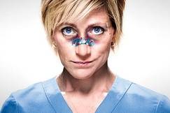 Edie Falco as Nurse Jackie, in scrubs with bandages on her nose, staring intensely ahead.