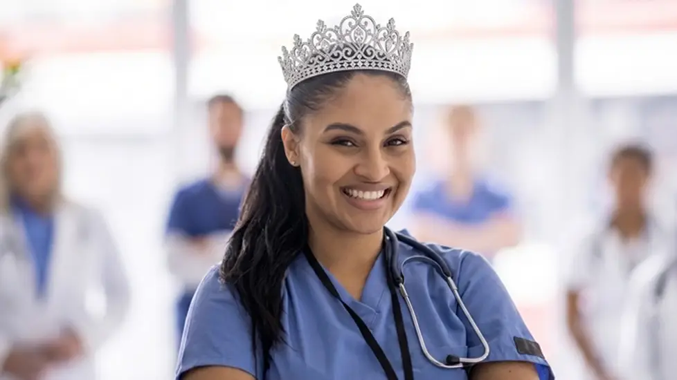 A portrait photograph shows a smiling nurse in blue scrubs wearing a stethoscope and an ornate silver tiara, with blurred hospital colleagues in the background.