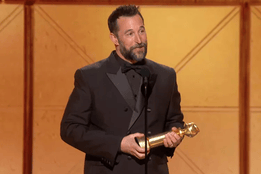 Noah Wyle in a dark tuxedo standing at a microphone at the Golden Globes awards stage, holding a statuette in both hands.