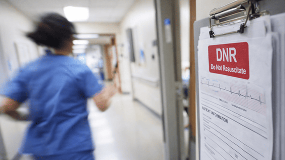 Nurse in scrubs rushing down a hospital hallway as a DNR medical chart is posted outside a patient room.