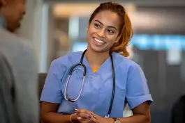Smiling nurse in blue scrubs with a stethoscope around her neck talking with a patient in a clinical setting.