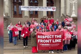 Group of nurses gathered on the steps of a government building holding signs and a large banner reading “Respect the Front Line,” advocating against mandatory overtime and for better working condition