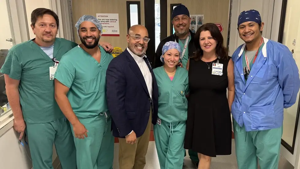 David Zambrana (navy blazer) with smiling nurses and clinicians in green scrubs in Jackson Memorial hallway.