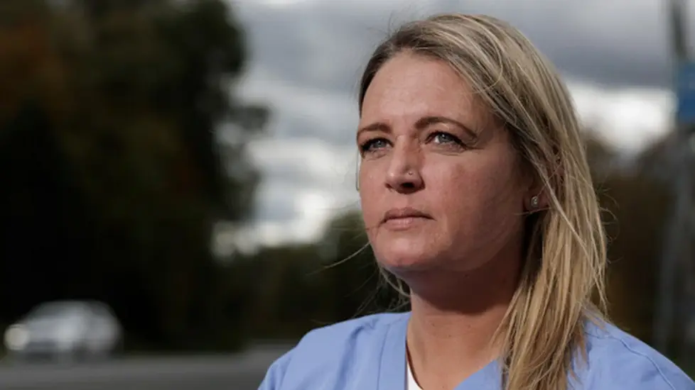 Andrea Elia wearing light blue medical scrubs sits outdoors near a roadside, looking thoughtful into the distance. The sky is cloudy, and blurred trees and a car appear in the background.