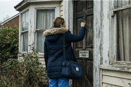 A home health nurse seen from behind knocks on the weathered door of a dilapidated house marked with a "PRIVATE PROPERTY - BEWARE" sign.