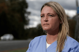 Andrea Elia wearing light blue medical scrubs sits outdoors near a roadside, looking thoughtful into the distance. The sky is cloudy, and blurred trees and a car appear in the background.