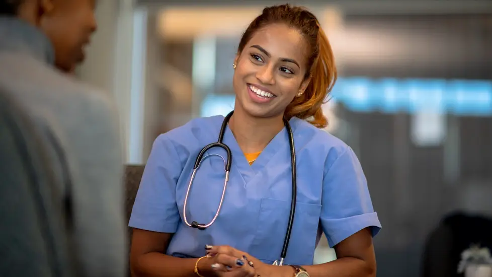 Smiling nurse in blue scrubs with a stethoscope around her neck talking with a patient in a clinical setting.