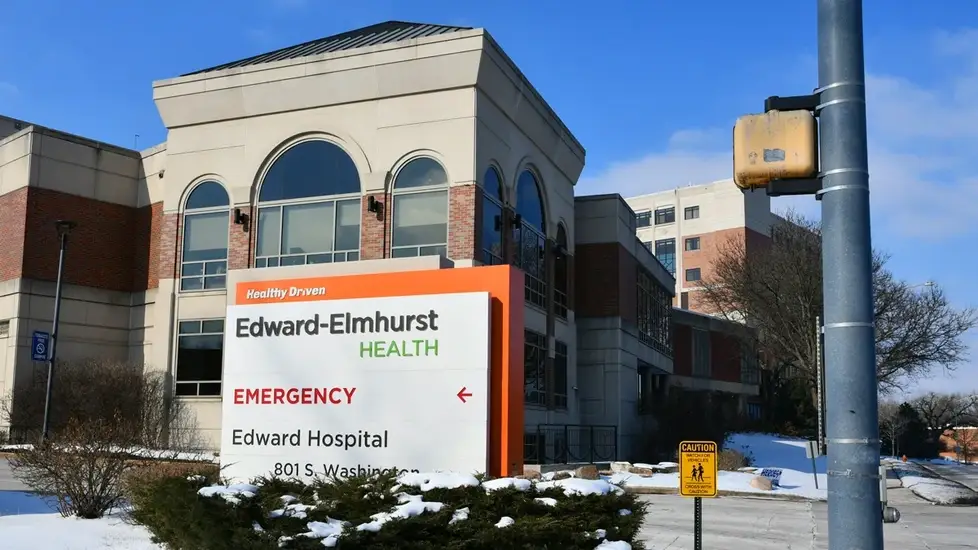 Front entrance of Edward Hospital in Naperville, Illinois, with a large Edward-Elmhurst Health sign and emergency directions, surrounded by snow-covered ground and bushes on a clear winter day.