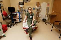 ER nurse Hannah Cvancara sitting on a hospital stretcher in an emergency room, wearing green scrubs and showing her prosthetic left leg.