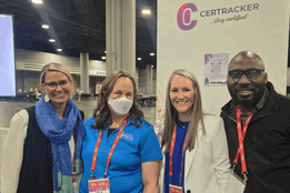 CerTracker team smiling at conference booth: two women in lanyards (one blue shirt, one white blazer), man in glasses and navy jacket. Purple logo backdrop.