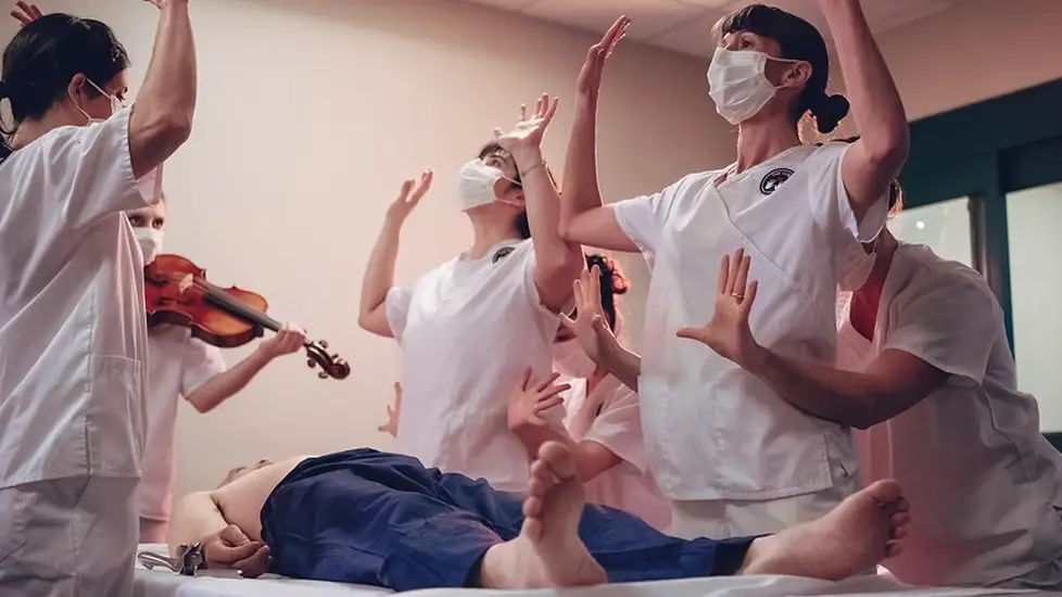 Nurses in white scrubs perform expressive dance movements around a patient on a hospital bed during Tara Rynders' "First Do No Harm."