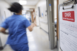 Nurse in scrubs rushing down a hospital hallway as a DNR medical chart is posted outside a patient room.