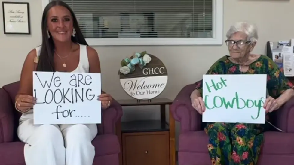 A nurse and an older woman sit holding signs that read “We are looking for…” and “Hot Cowboys” at Gainesville Health Care Center.