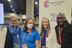 CerTracker team smiling at conference booth: two women in lanyards (one blue shirt, one white blazer), man in glasses and navy jacket. Purple logo backdrop.