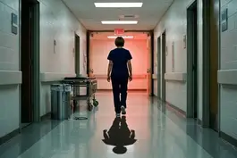 A nurse in scrubs walks toward a glowing "EXIT" sign in a long, sterile hospital hallway. On the left, her discarded stethoscope hangs over a trash can near a gurney.