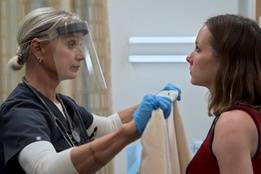 The Pitt's Sexual Assault Nurse Examiner Dana Evans wearing protective gear prepares evidence collection materials while speaking with a patient in an exam room.