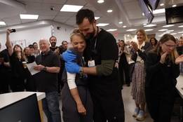 Noah Wyle and Katherine LaNasa embrace, celebrating The Pitt's 13 Emmy nominations while the cast and crew, dressed in hospital scrubs, gather in a brightly lit breakroom to cheer them on.