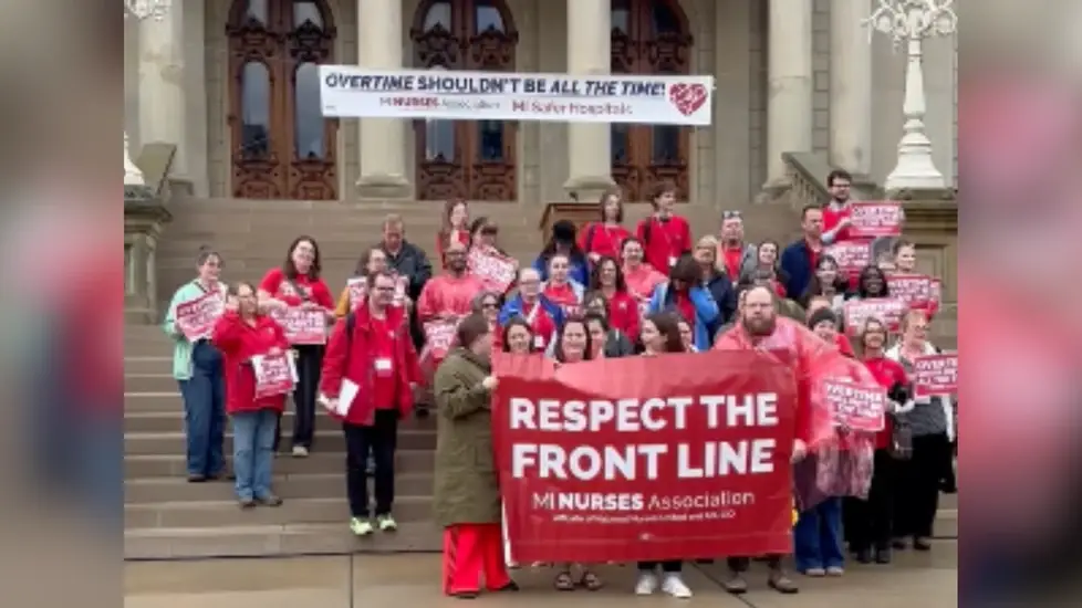 Group of nurses gathered on the steps of a government building holding signs and a large banner reading “Respect the Front Line,” advocating against mandatory overtime and for better working condition