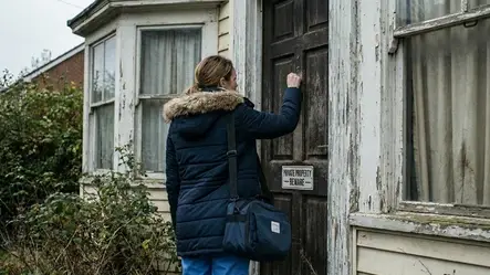 A home health nurse seen from behind knocks on the weathered door of a dilapidated house marked with a "PRIVATE PROPERTY - BEWARE" sign.