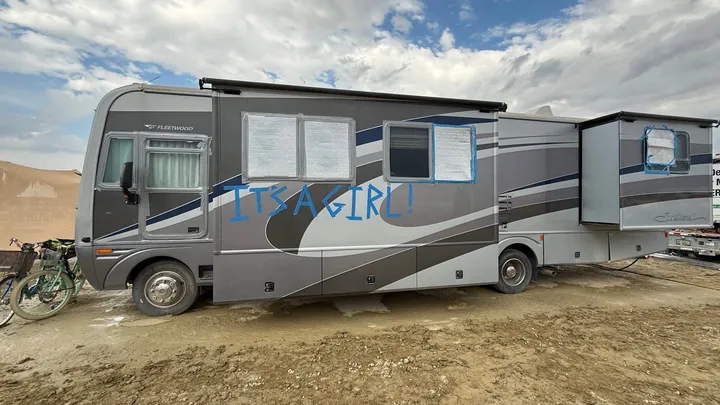Gray RV parked at Burning Man with “IT’S A GIRL!” painted in blue across the side, marking the surprise birth of baby Aurora.