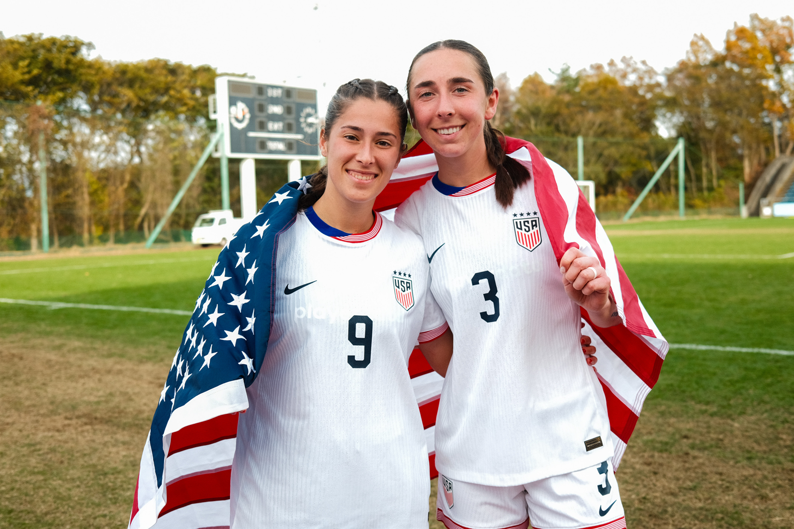 Paige Beaudry and a teammate stand on a soccer field draped in U.S. flags, wearing white USA jerseys after a game.